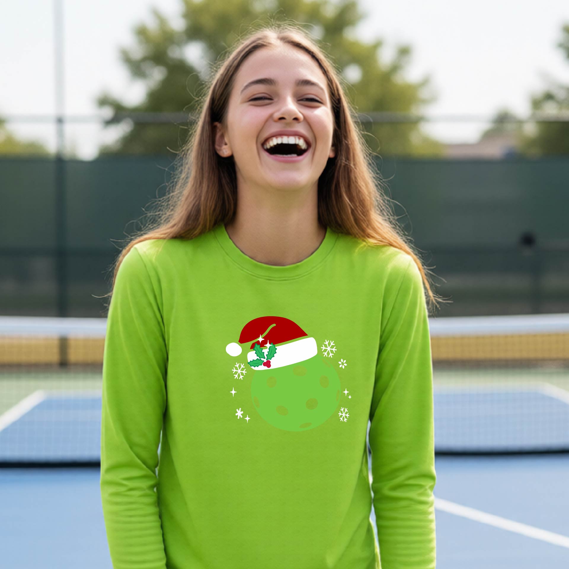 A young person laughing on an outdoor pickleball court wearing a lime green long-sleeve shirt with a green polka-dot pickleball graphic wearing a red Santa hat.