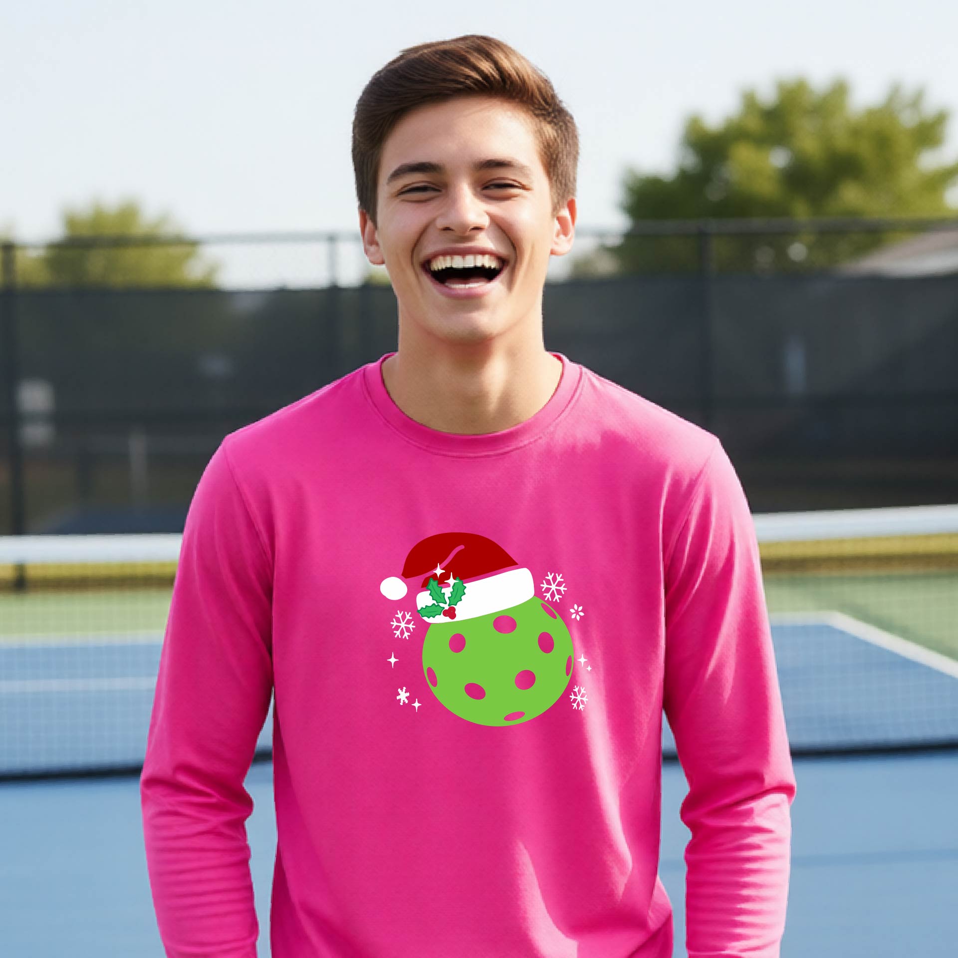 A young person laughing on an outdoor pickleball court wearing a pink long-sleeve shirt with a green polka-dot pickleball graphic wearing a red Santa hat.
