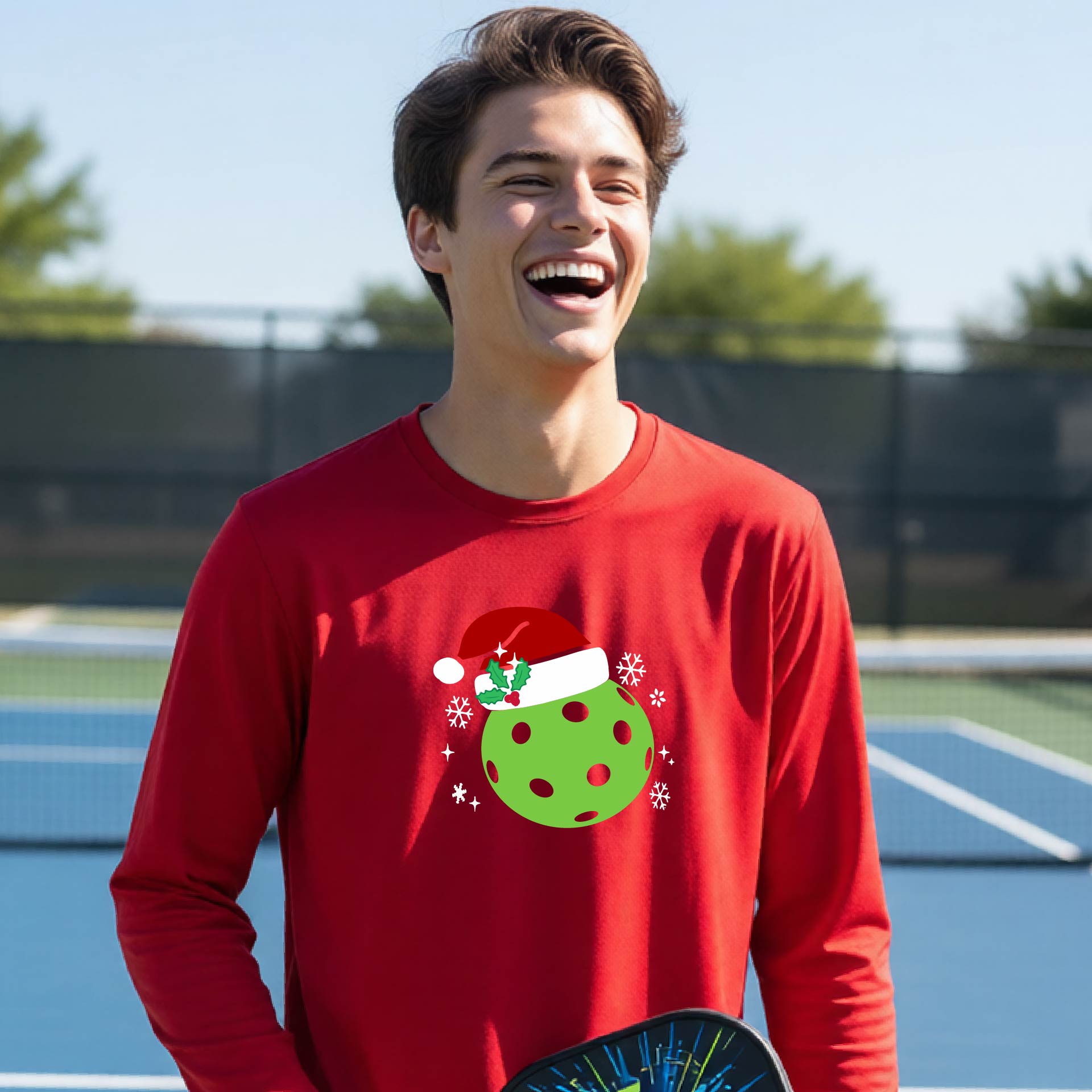 A young person laughing on an outdoor pickleball court wearing a red long-sleeve shirt with a green polka-dot pickleball graphic wearing a red Santa hat.