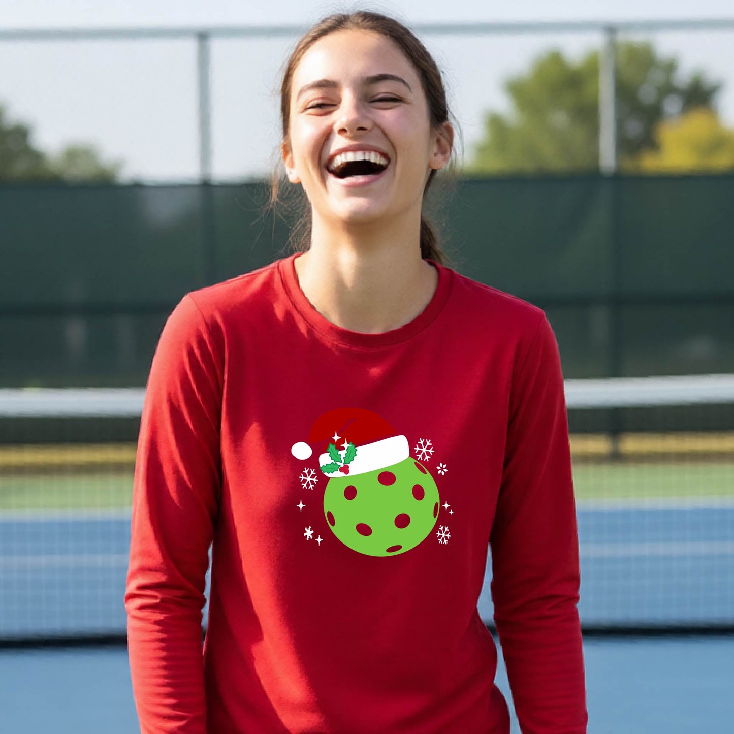 A young person laughing on an outdoor pickleball court wearing a red long-sleeve shirt with a green polka-dot pickleball graphic wearing a red Santa hat.