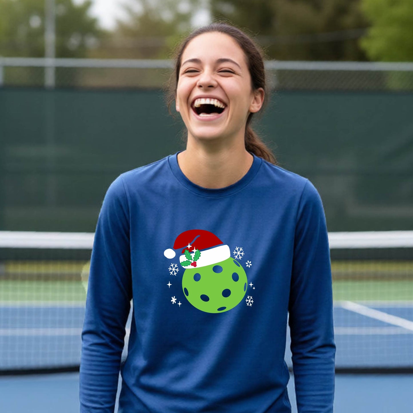A young person laughing on an outdoor pickleball court wearing a blue long-sleeve shirt with a green polka-dot pickleball graphic wearing a red Santa hat.