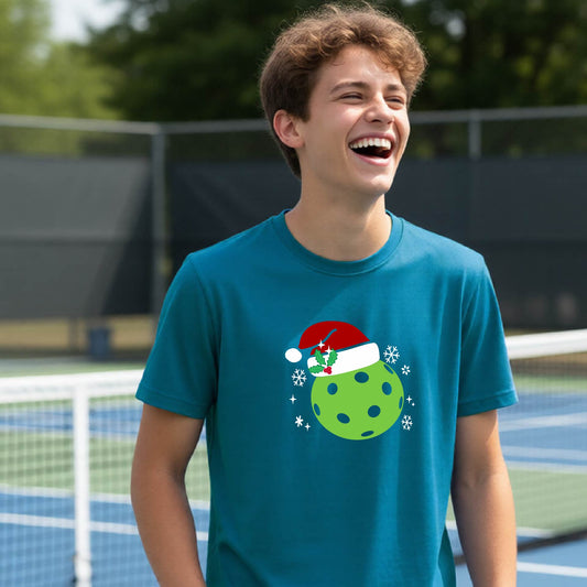 A smiling person standing on a blue pickleball court wearing a teal T-shirt with a festive green pickleball graphic wearing a red Santa hat.