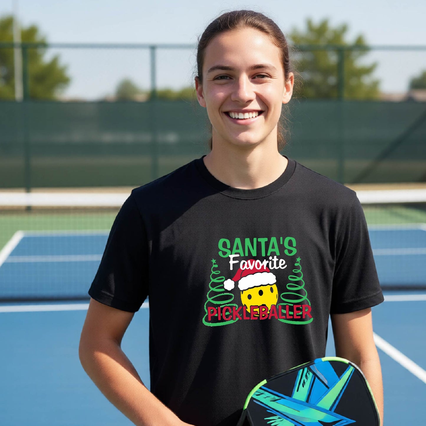 A young man laughing on an outdoor pickleball court, wearing a black t-shirt with a festive graphic that reads 'Santa's Favorite Pickleballer' with a yellow pickle, green pine trees, and a white Christmas hat.