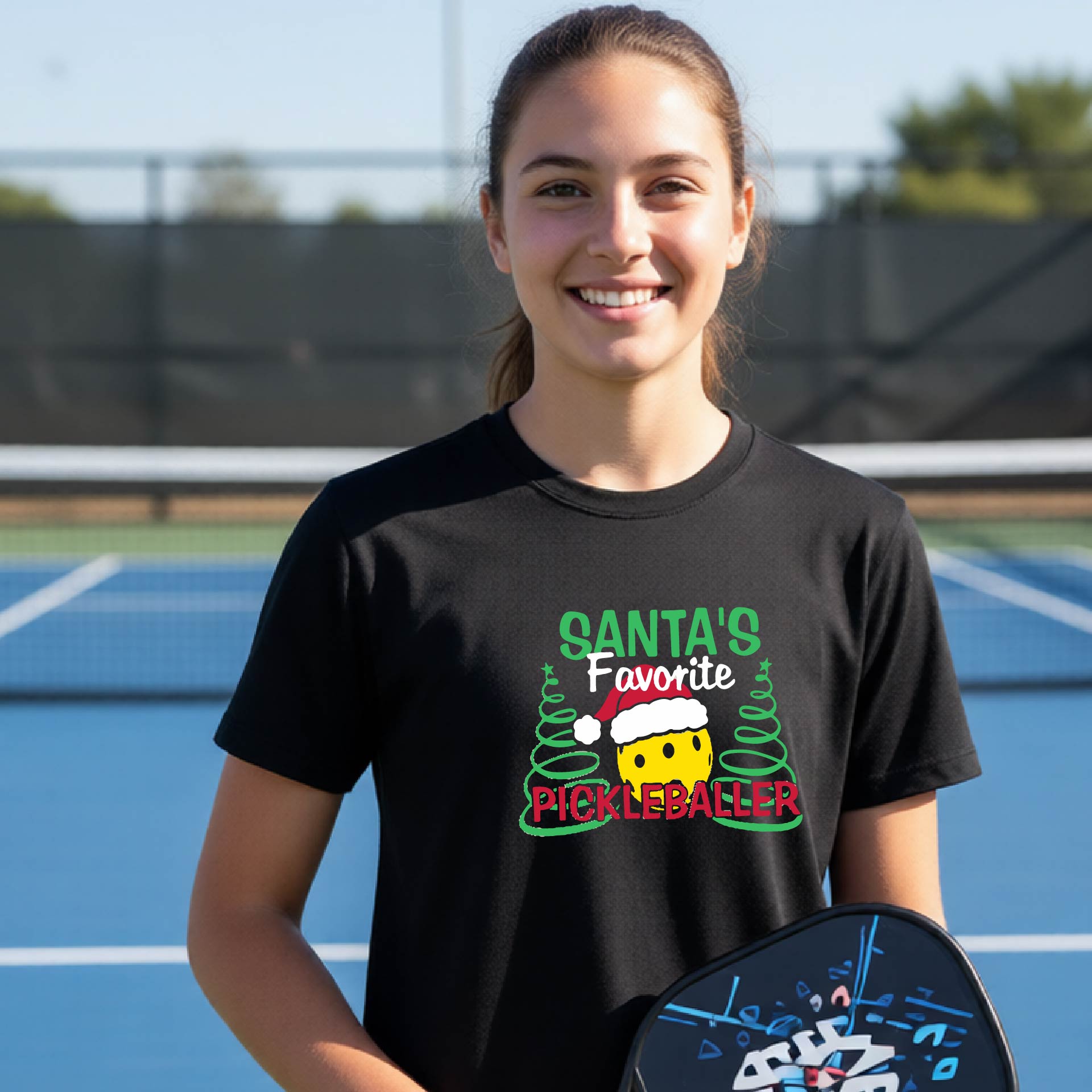 A young woman laughing on an outdoor pickleball court, wearing a black t-shirt with a festive graphic that reads 'Santa's Favorite Pickleballer' with a yellow pickle, green pine trees, and a white Christmas hat.
