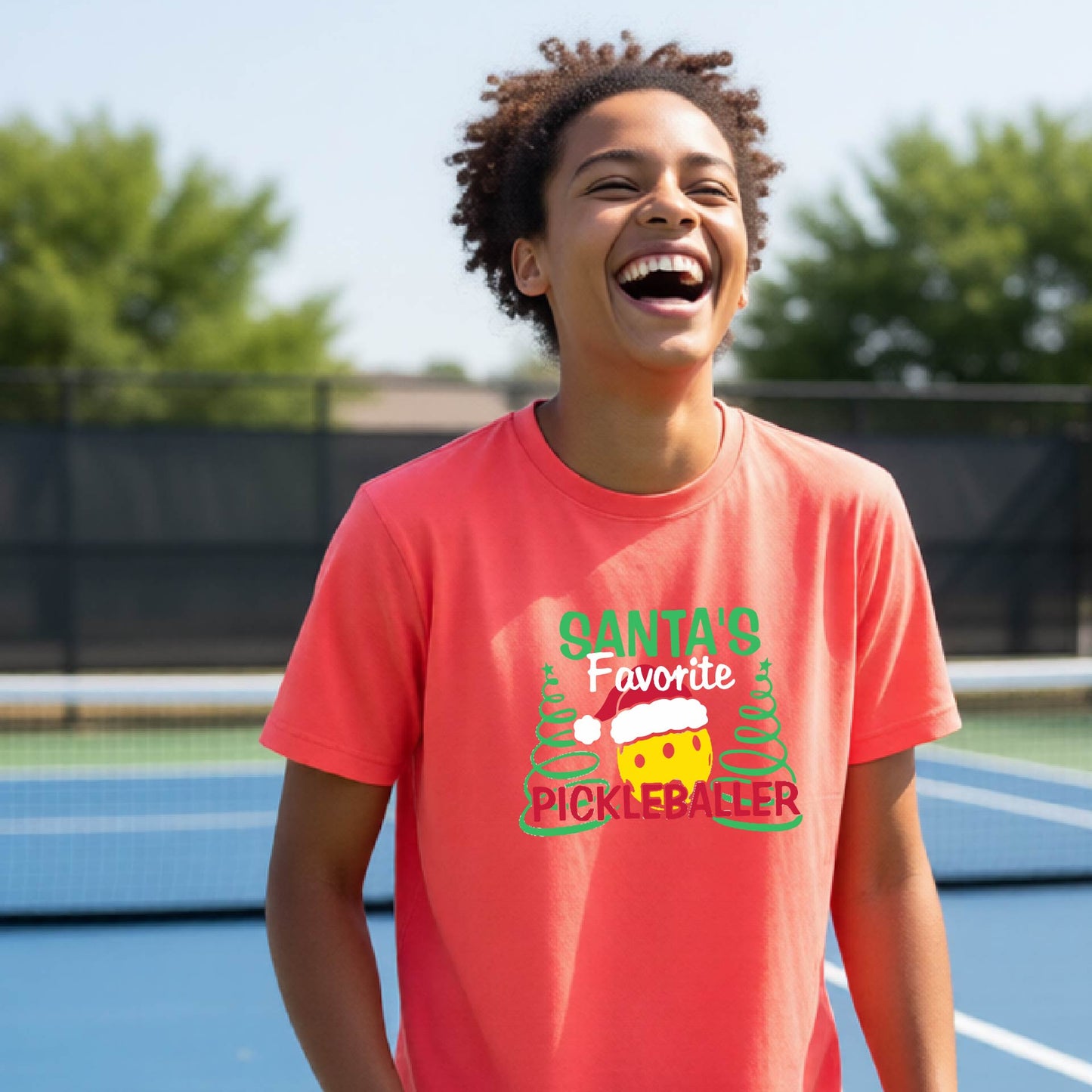 A young man laughing on an outdoor pickleball court, wearing a coral-colored  t-shirt with a festive graphic that reads 'Santa's Favorite Pickleballer' with a yellow pickle, green pine trees, and a white Christmas hat.