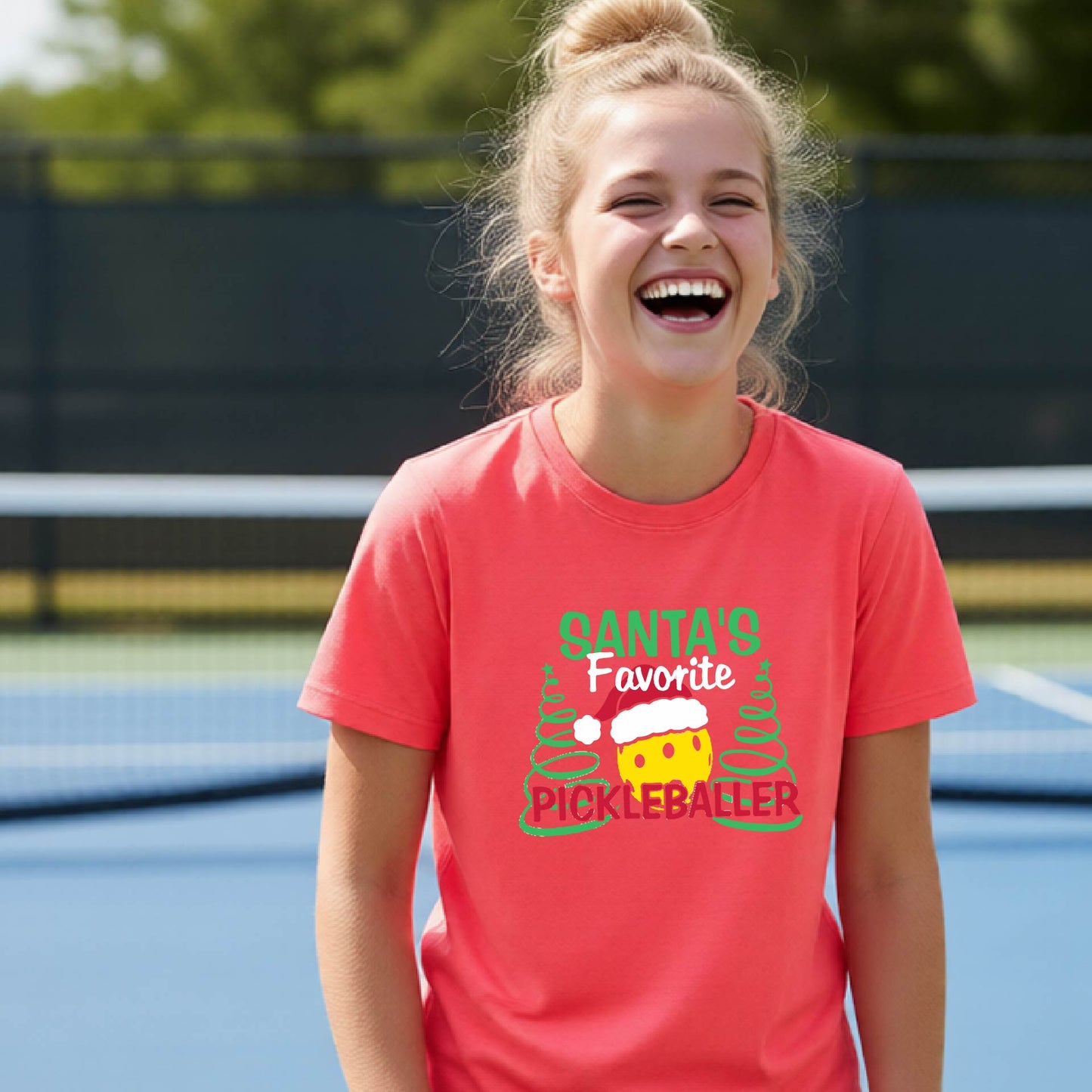 A young woman laughing on an outdoor pickleball court, wearing a coral-colored t-shirt with a festive graphic that reads 'Santa's Favorite Pickleballer' with a yellow pickle, green pine trees, and a white Christmas hat.