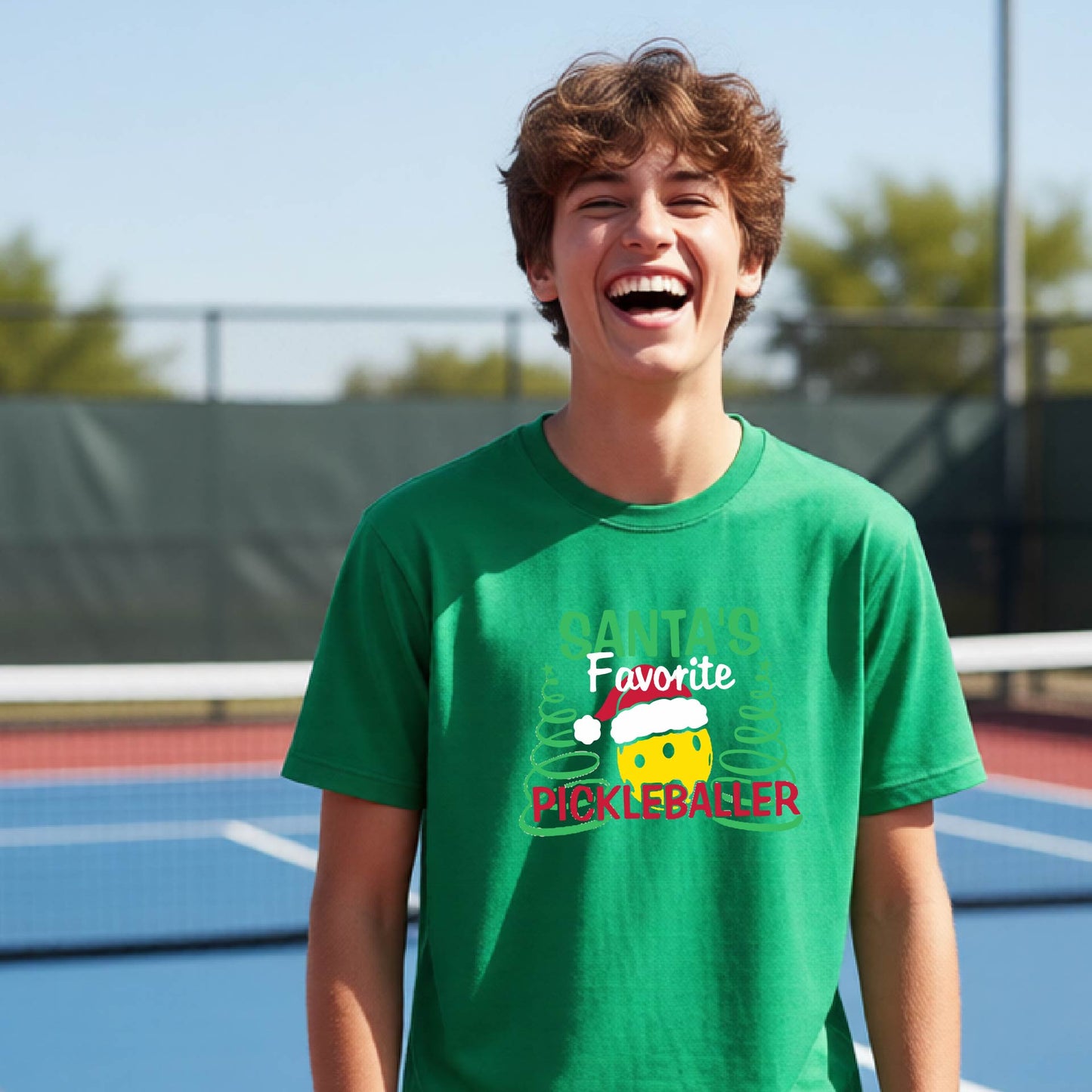A young man laughing on an outdoor pickleball court, wearing a green t-shirt with a festive graphic that reads 'Santa's Favorite Pickleballer' with a yellow pickle, green pine trees, and a white Christmas hat.