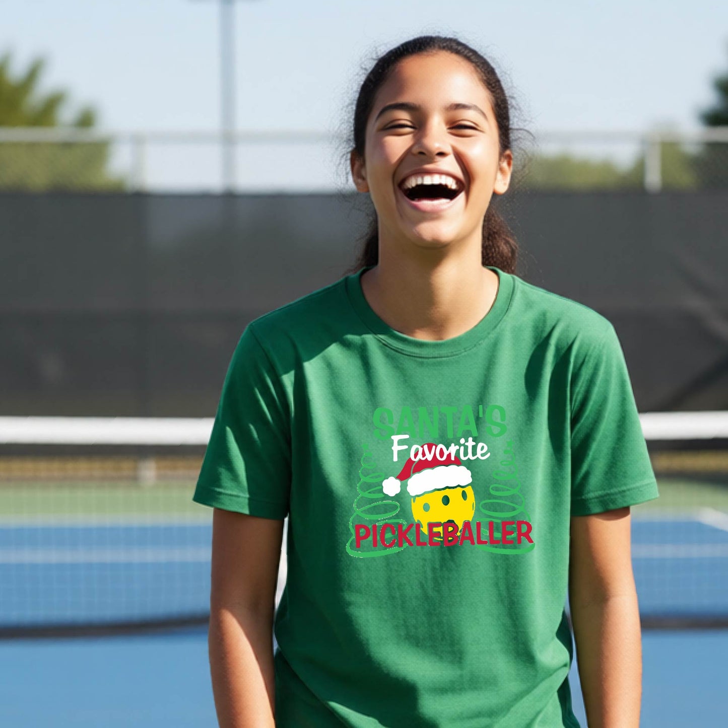 A young woman laughing on an outdoor pickleball court, wearing a green t-shirt with a festive graphic that reads 'Santa's Favorite Pickleballer' with a yellow pickle, green pine trees, and a white Christmas hat.