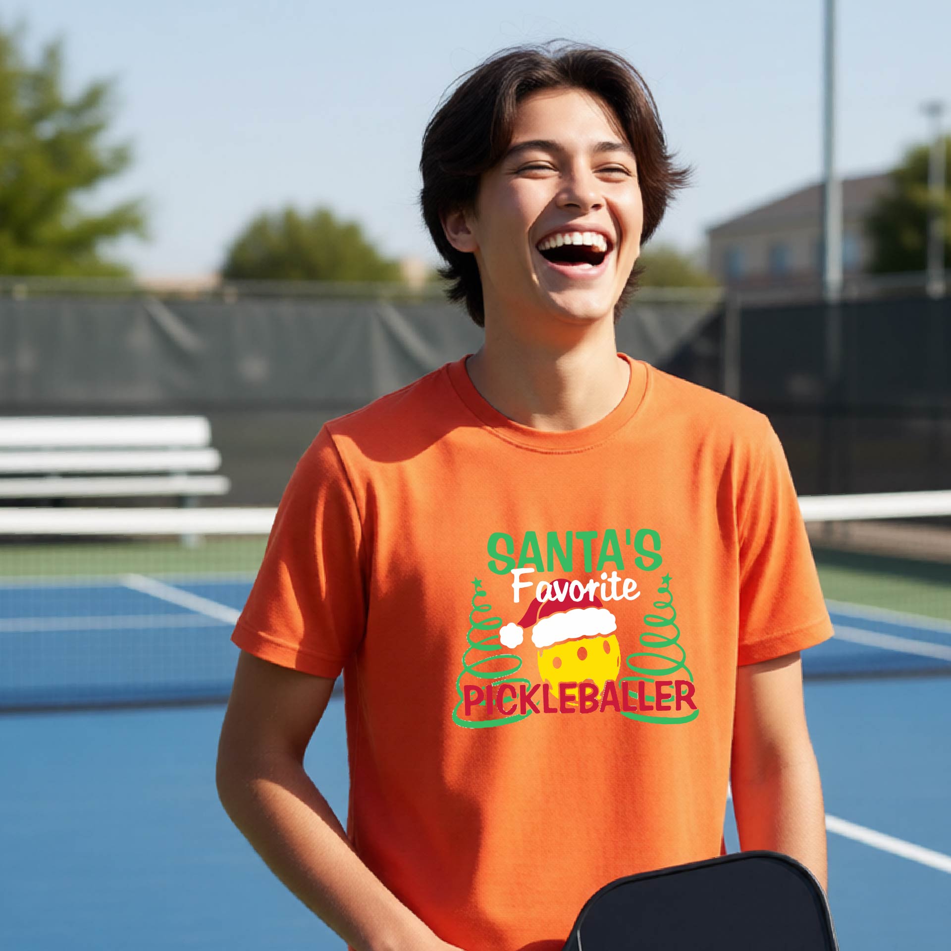 A young man laughing on an outdoor pickleball court, wearing an orange t-shirt with a festive graphic that reads 'Santa's Favorite Pickleballer' with a yellow pickle, green pine trees, and a white Christmas hat.