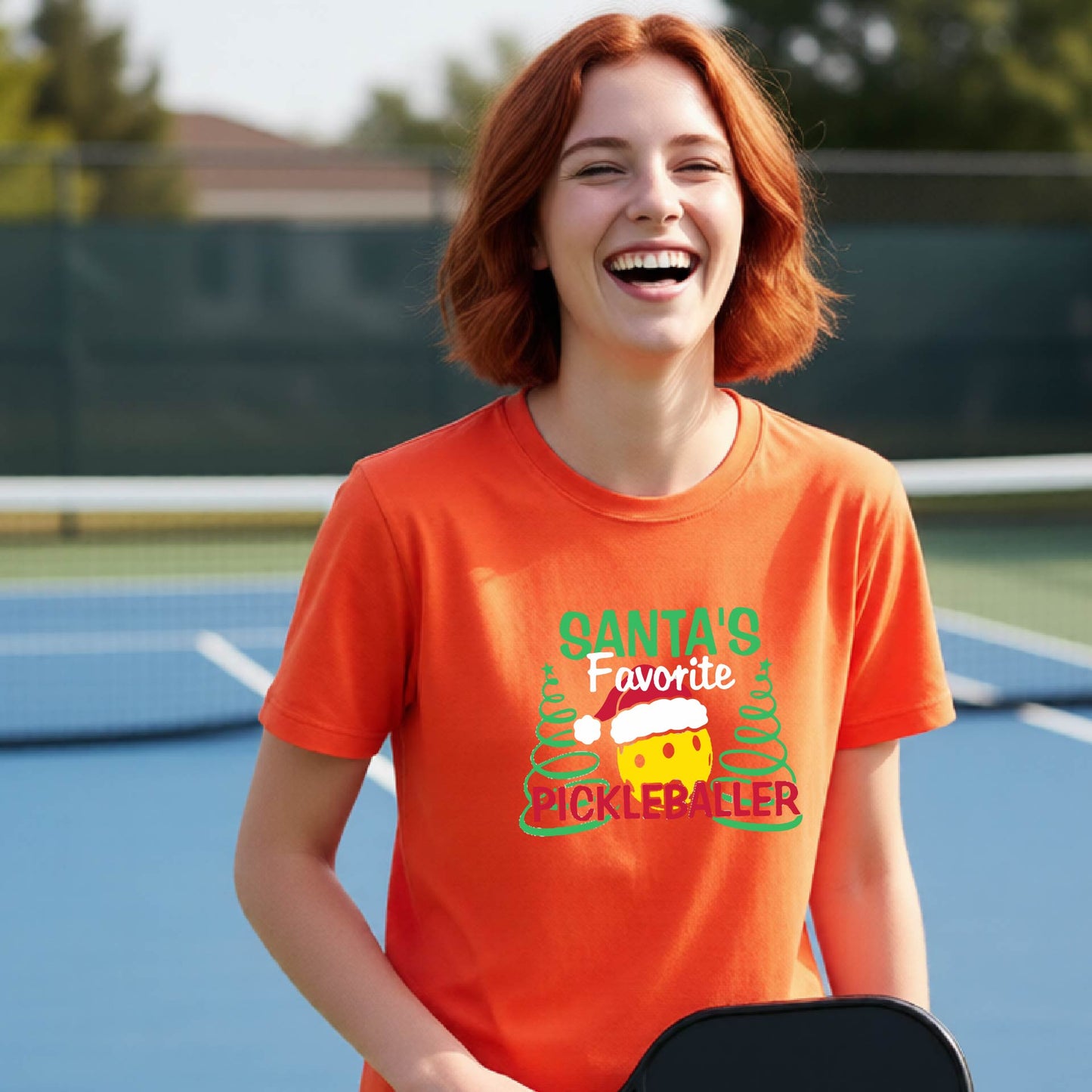 A young woman laughing on an outdoor pickleball court, wearing a orange t-shirt with a festive graphic that reads 'Santa's Favorite Pickleballer' with a yellow pickle, green pine trees, and a white Christmas hat.