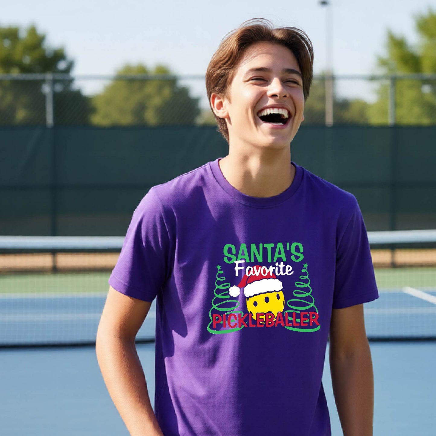 A young man laughing on an outdoor pickleball court, wearing a purple t-shirt with a festive graphic that reads 'Santa's Favorite Pickleballer' with a yellow pickle, green pine trees, and a white Christmas hat.