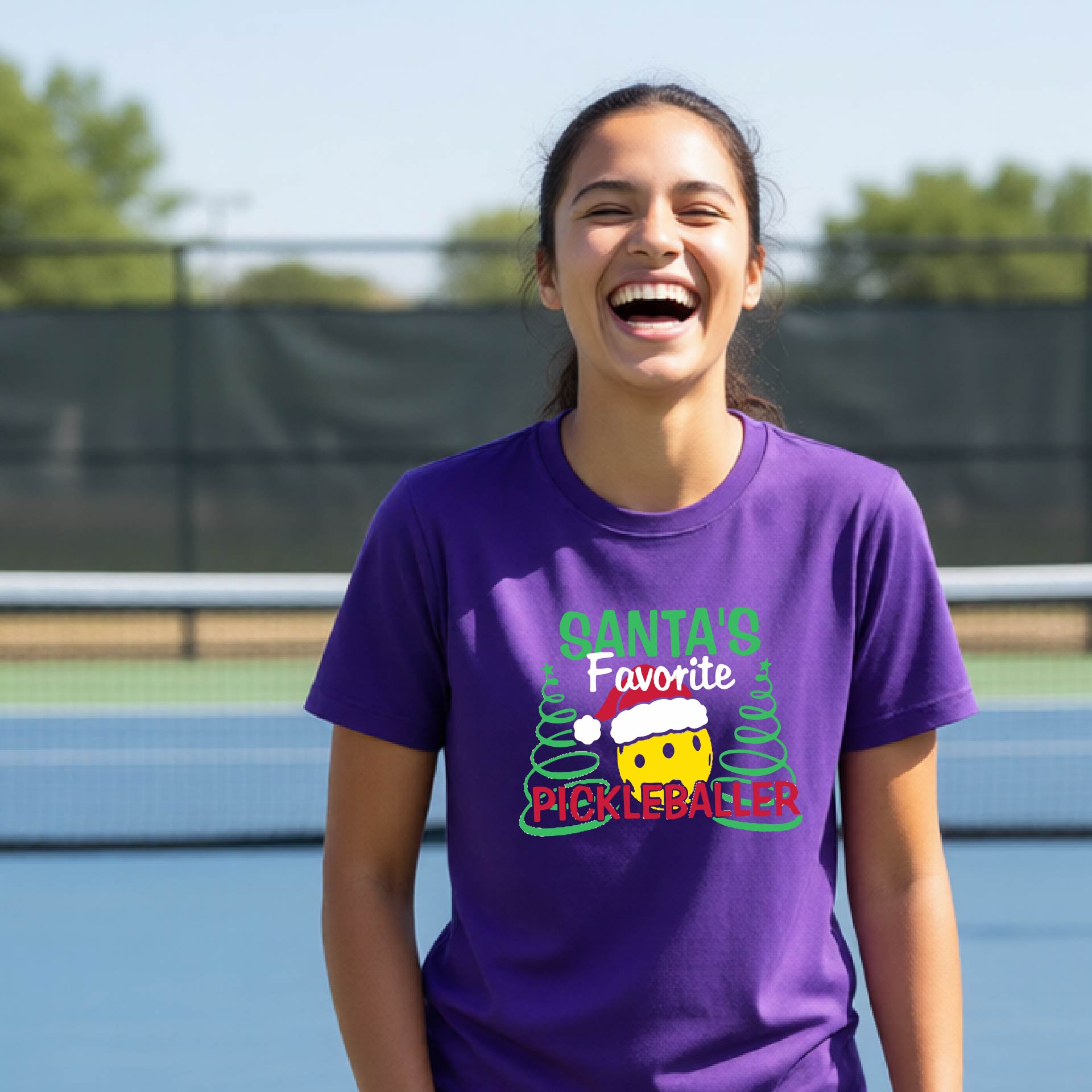 A young woman laughing on an outdoor pickleball court, wearing a purple t-shirt with a festive graphic that reads 'Santa's Favorite Pickleballer' with a yellow pickle, green pine trees, and a white Christmas hat.