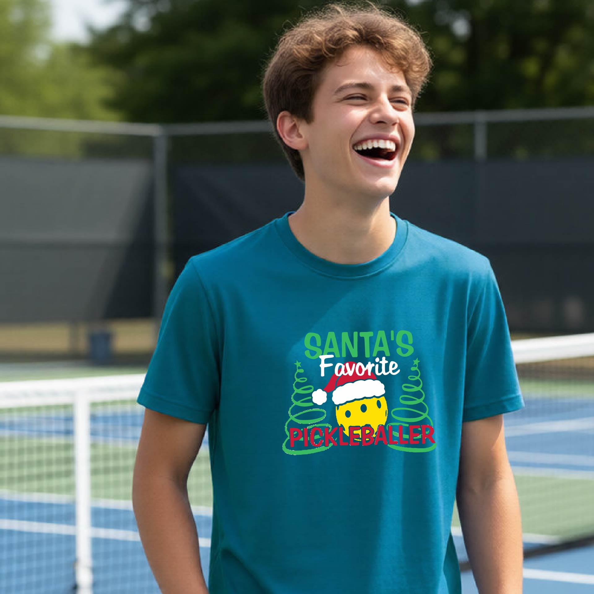 A young man laughing on an outdoor pickleball court, wearing a teal t-shirt with a festive graphic that reads 'Santa's Favorite Pickleballer' with a yellow pickle, green pine trees, and a white Christmas hat.