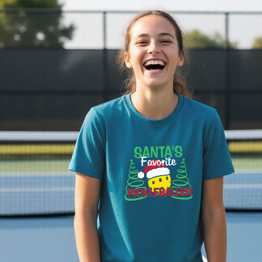 A young woman laughing on an outdoor pickleball court, wearing a teal t-shirt with a festive graphic that reads 'Santa's Favorite Pickleballer' with a yellow pickle, green pine trees, and a white Christmas hat.