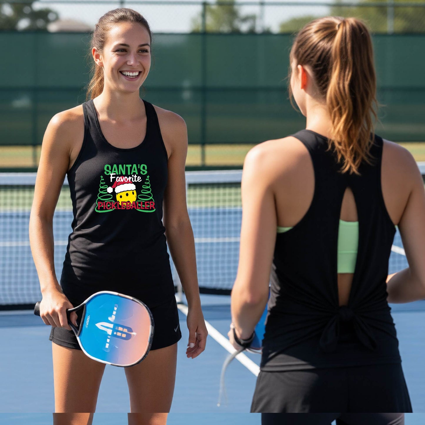 Two women on a pickleball court wearing matching black tank tops. The woman on the left faces the camera and wears a tank top with a colorful graphic that reads 'Santa's Favorite Pickleballer' with a candy cane and pine trees.