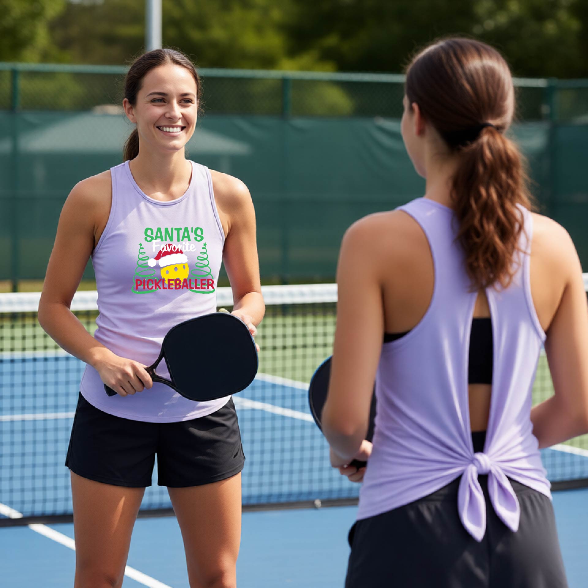 Two women on a pickleball court wearing matching light lavender tank tops. The woman on the left faces the camera and wears a tank top with a colorful graphic that reads 'Santa's Favorite Pickleballer' with a candy cane and pine trees.
