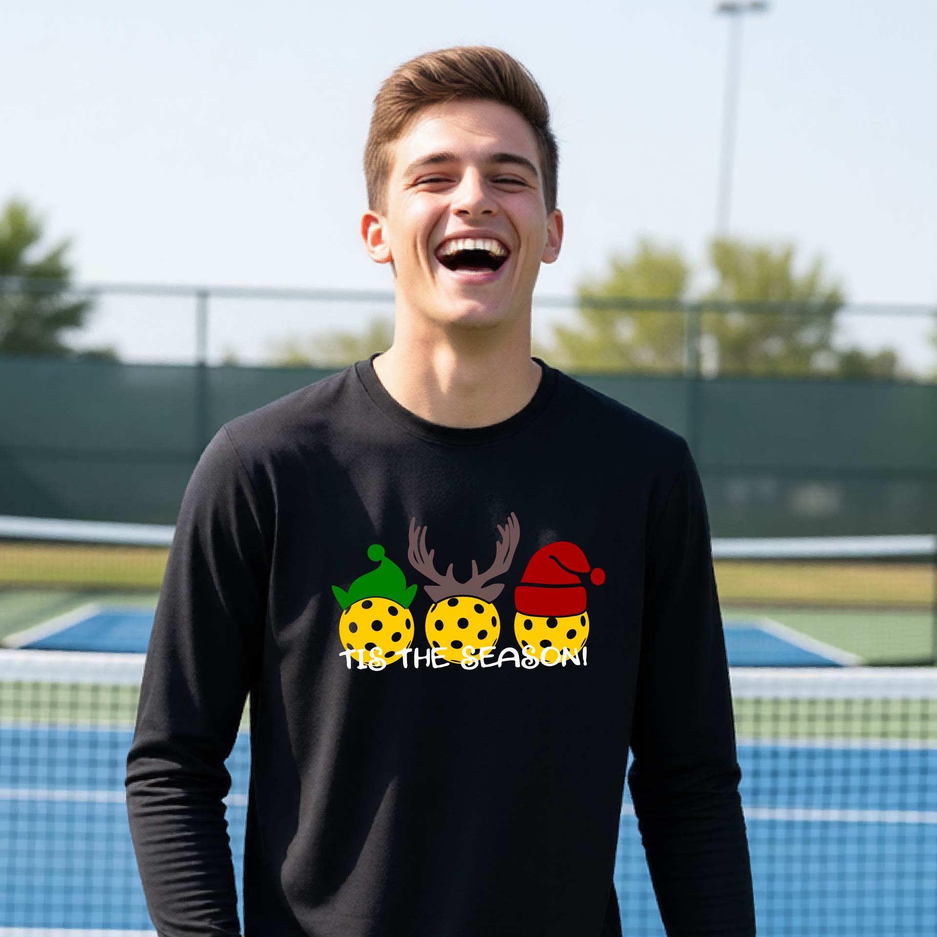 A smiling young person on a blue pickleball court wearing a lime green t-shirt with 'TIS THE SEASON' printed in white, with pickleballs decorated wearing antlers and Christmas hats. 