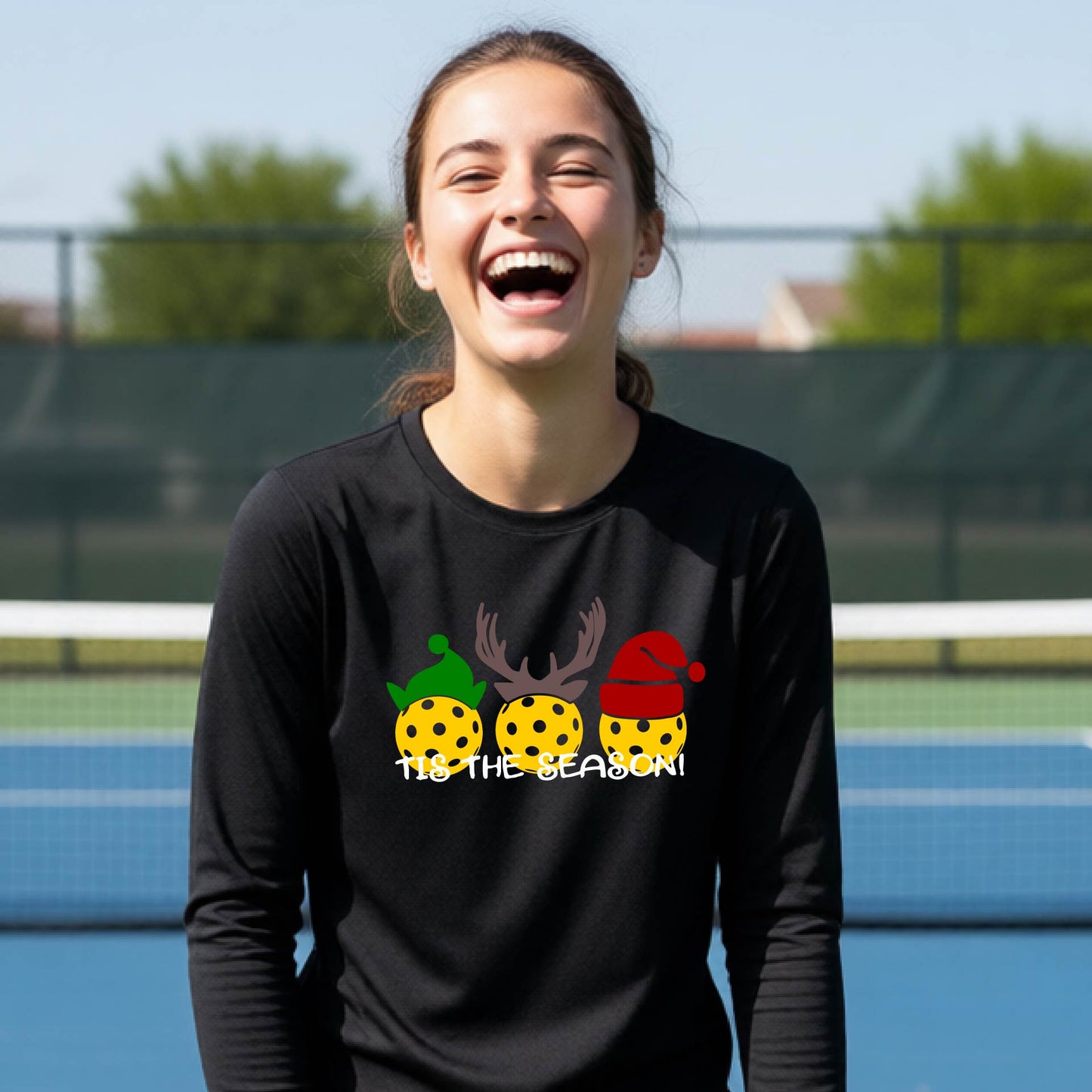 A smiling young person on a blue pickleball court wearing a black t-shirt with 'TIS THE SEASON' printed in white, with pickleballs decorated wearing antlers and Christmas hats. 
