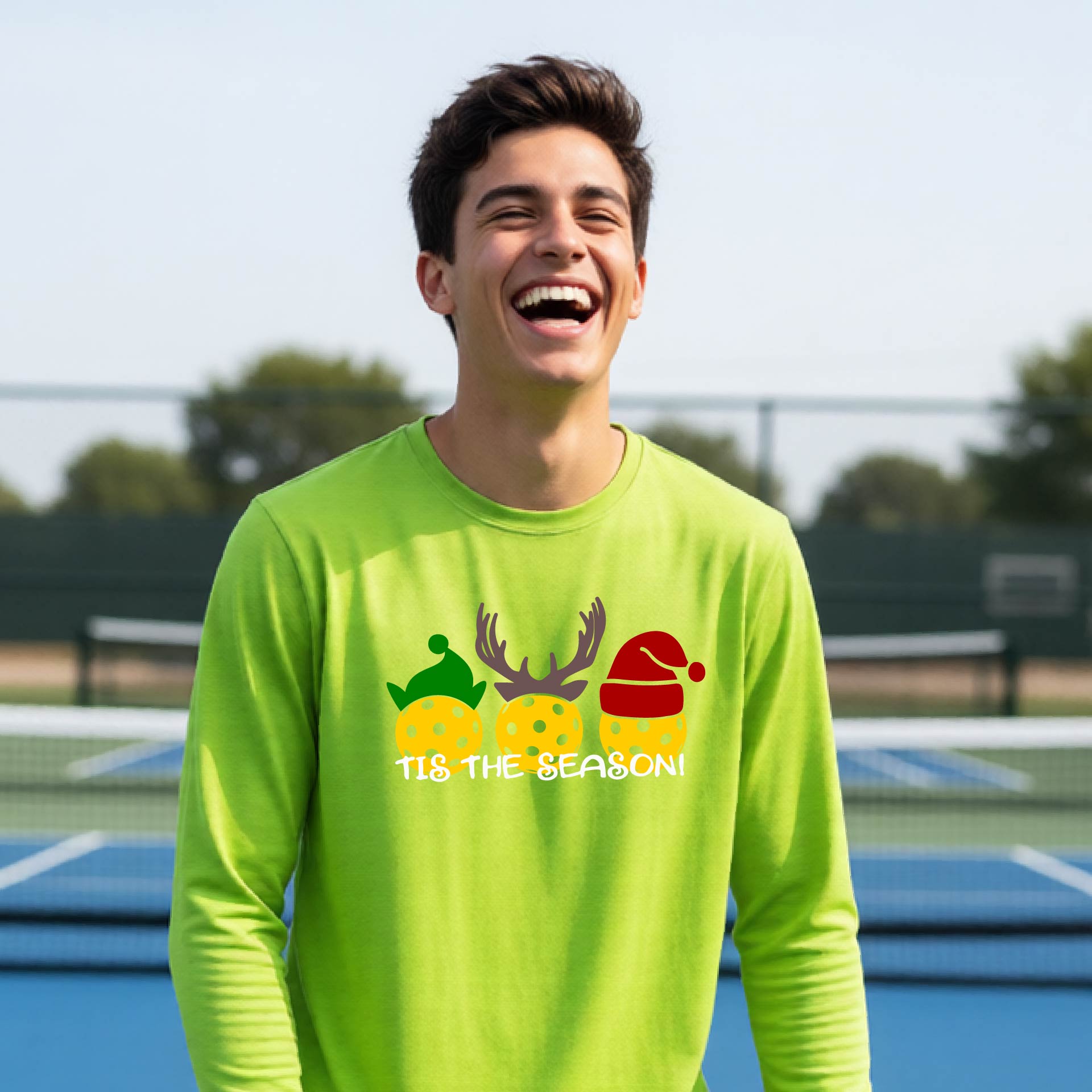 A smiling young person on a blue pickleball court wearing a lime green t-shirt with 'TIS THE SEASON' printed in white, with pickleballs decorated wearing antlers and Christmas hats. 