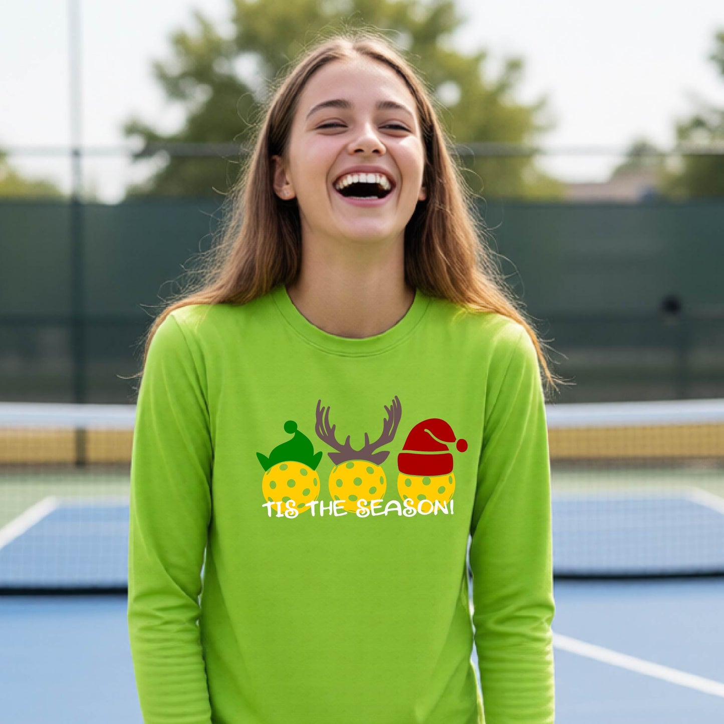 A smiling young person on a blue pickleball court wearing a lime green t-shirt with 'TIS THE SEASON' printed in white, with pickleballs decorated wearing antlers and Christmas hats. 