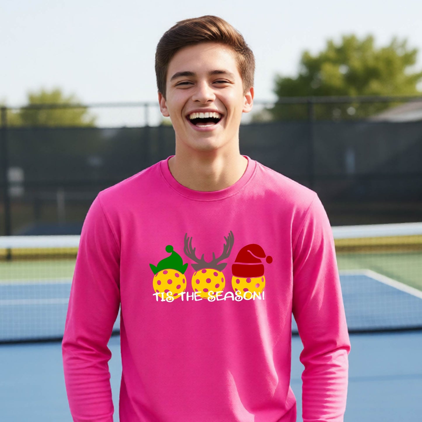 A smiling young person on a blue pickleball court wearing a bright pink t-shirt with 'TIS THE SEASON' printed in white, with pickleballs decorated wearing antlers and Christmas hats. 