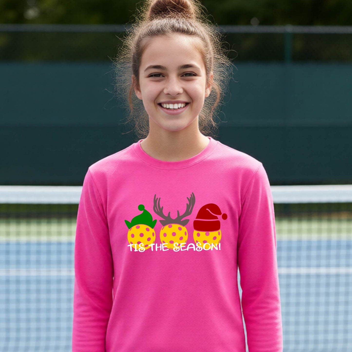 A smiling young person on a blue pickleball court wearing a pink t-shirt with 'TIS THE SEASON' printed in white, with pickleballs decorated wearing antlers and Christmas hats. 