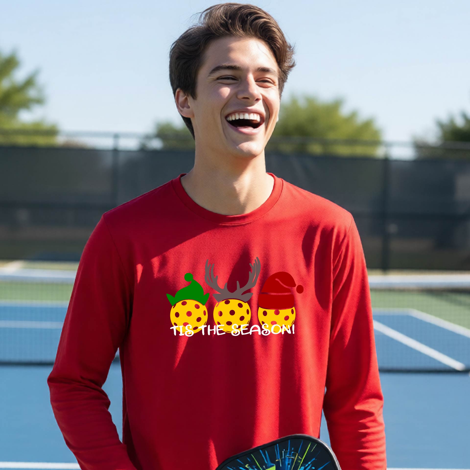 A smiling young person on a blue pickleball court wearing a red t-shirt with 'TIS THE SEASON' printed in white, with pickleballs decorated wearing antlers and Christmas hats. 
