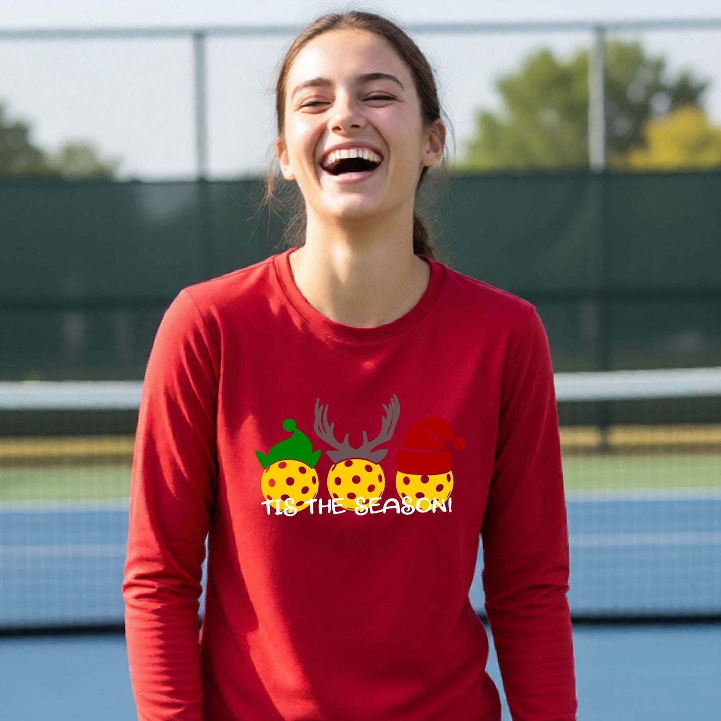 A smiling young person on a blue pickleball court wearing a bright red t-shirt with 'TIS THE SEASON' printed in white, with pickleballs decorated wearing antlers and Christmas hats. 