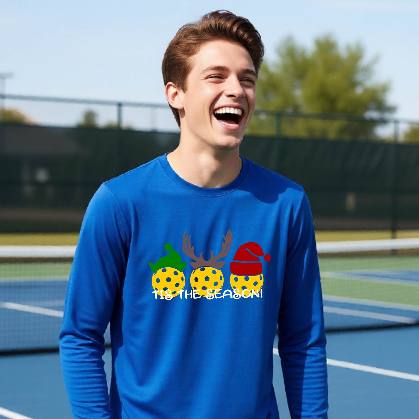 A smiling young person on a blue pickleball court wearing a royal blue t-shi rt with 'TIS THE SEASON' printed in white, with pickleballs decorated wearing antlers and Christmas hats. 