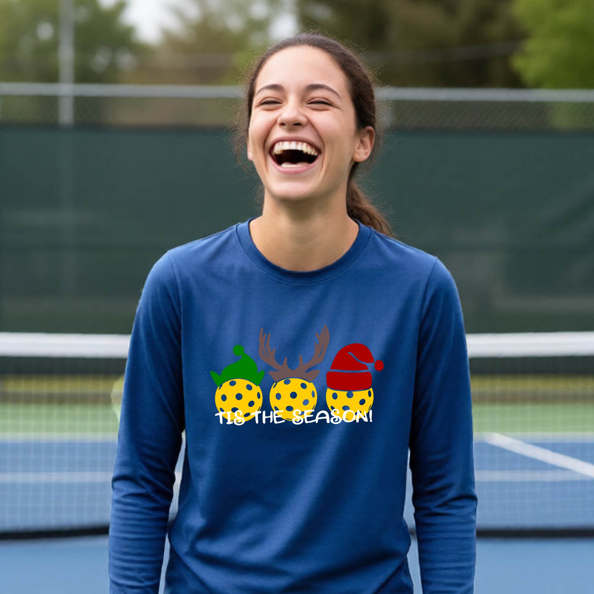 A smiling young person on a blue pickleball court wearing a royal  blue t-shirt with 'TIS THE SEASON' printed in white, with pickleballs decorated wearing antlers and Christmas hats. 