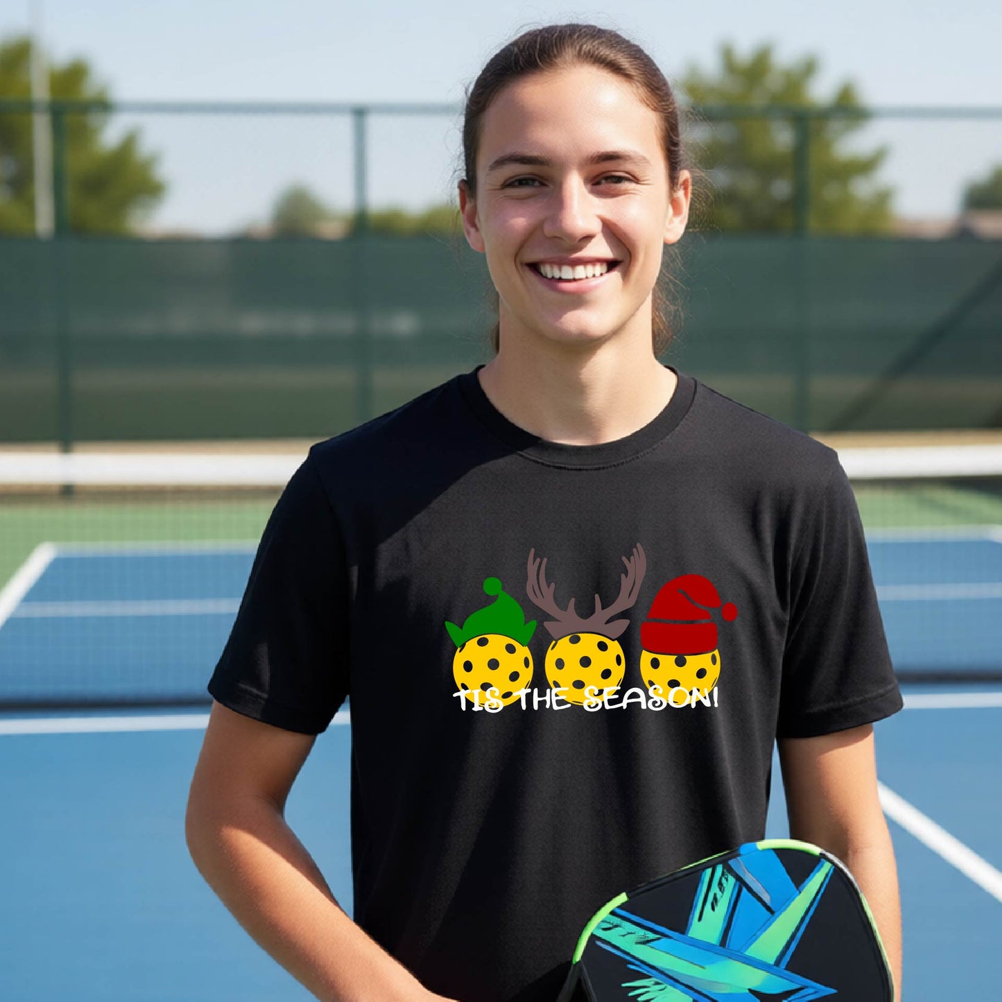 A smiling woman on a blue pickleball court wearing a black graphic t-shirt with a Christmas-themed design of three  pickleballs under antlers and a Santa hat, holding a paddle in her hand.