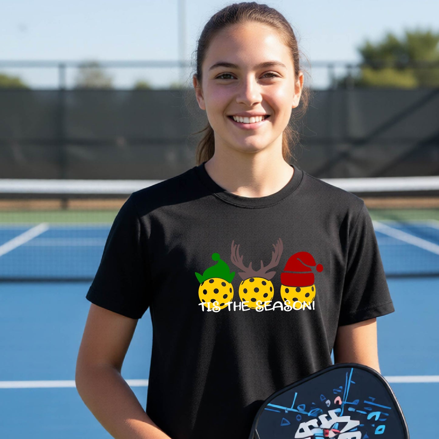 A smiling young woman stands on a blue pickleball court wearing a black graphic t-shirt with a holiday-themed design of three yellow pickleballs in green, brown, and red hats, with the text 'TIS THE SEASON!'. She is holding a black pickleball paddle.