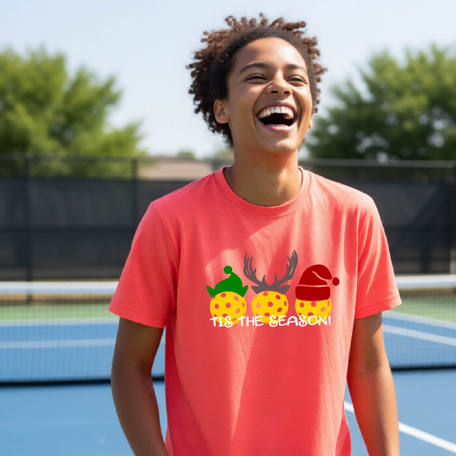 A person laughing on an outdoor pickleball court wearing a coral-pink t-shirt with a festive graphic of three pickleballs in holiday hats and the text '11.8 THE SEASON!'.