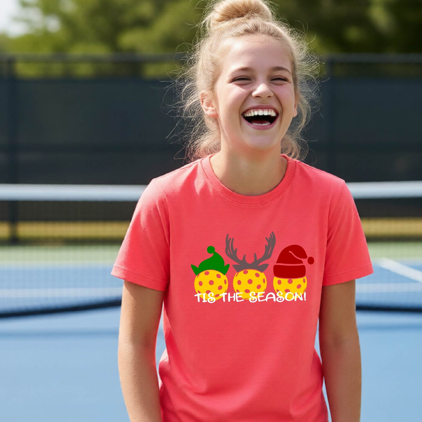 A young girl with her hair in a bun is laughing on an outdoor tennis court, wearing a coral pink t-shirt with a pickleball Christmas-themed graphic of three yellow ornaments and the text 'TIS THE SEASON!'.