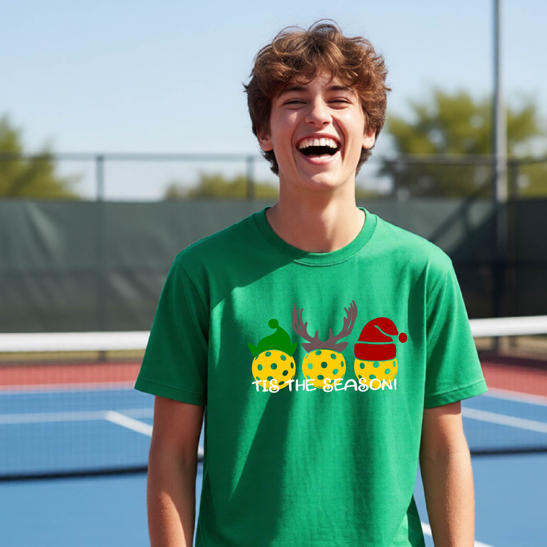 A young man laughing on an outdoor pickleball court wearing a green t-shirt with a Christmas-themed graphic of three pickleballs and the text 'IT'S THE SEASON!'.