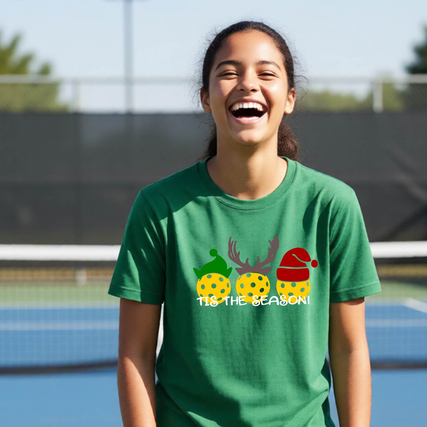 A woman laughing on an outdoor pickleball court wearing a green t-shirt with a festive Christmas-themed graphic of three yellow patterned pickleballs wearing Santa hats and the text 'TIS THE SEASON!'.