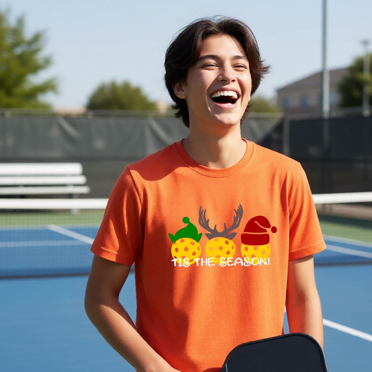 A young man laughing on a pickleball court wearing an orange t-shirt with a Christmas-themed graphic of three yellow pickleballs wearing holiday hats and the text 'TIS THE SEASON!'.