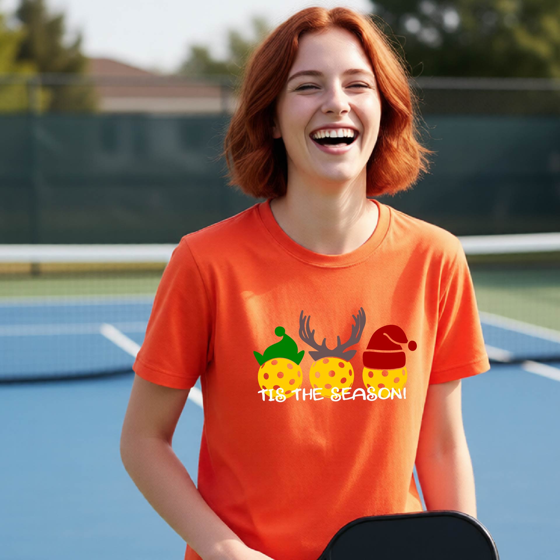 A smiling woman with red hair stands on a blue pickleball court wearing an orange graphic T-shirt. The shirt features three yellow pickleballs decorated with Christmas hats and antlers, with the text 'TIS THE SEASON!' printed below.