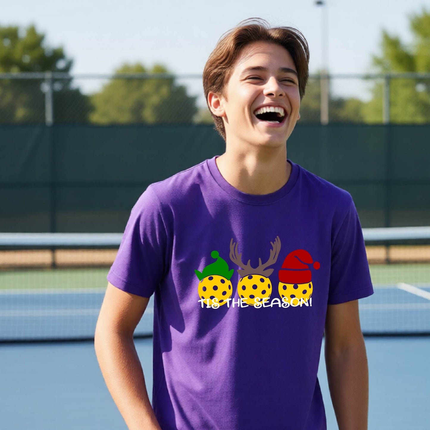 A man laughing on an outdoor pickleball court wearing a purple t-shirt with a graphic of three yellow pickleballs  and the text 'IT'S THE SEASON!'.