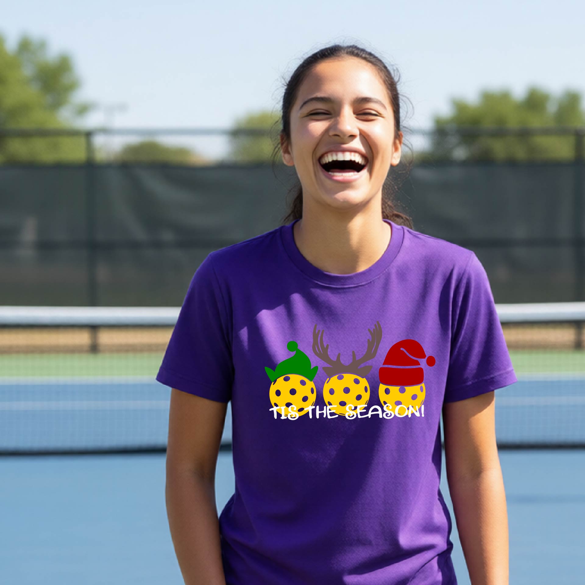 A woman laughing on an outdoor pickleball court wearing a purple t-shirt with a Christmas-themed graphic of three decorated pickleballs and the text 'IT'S THE SEASON!'.