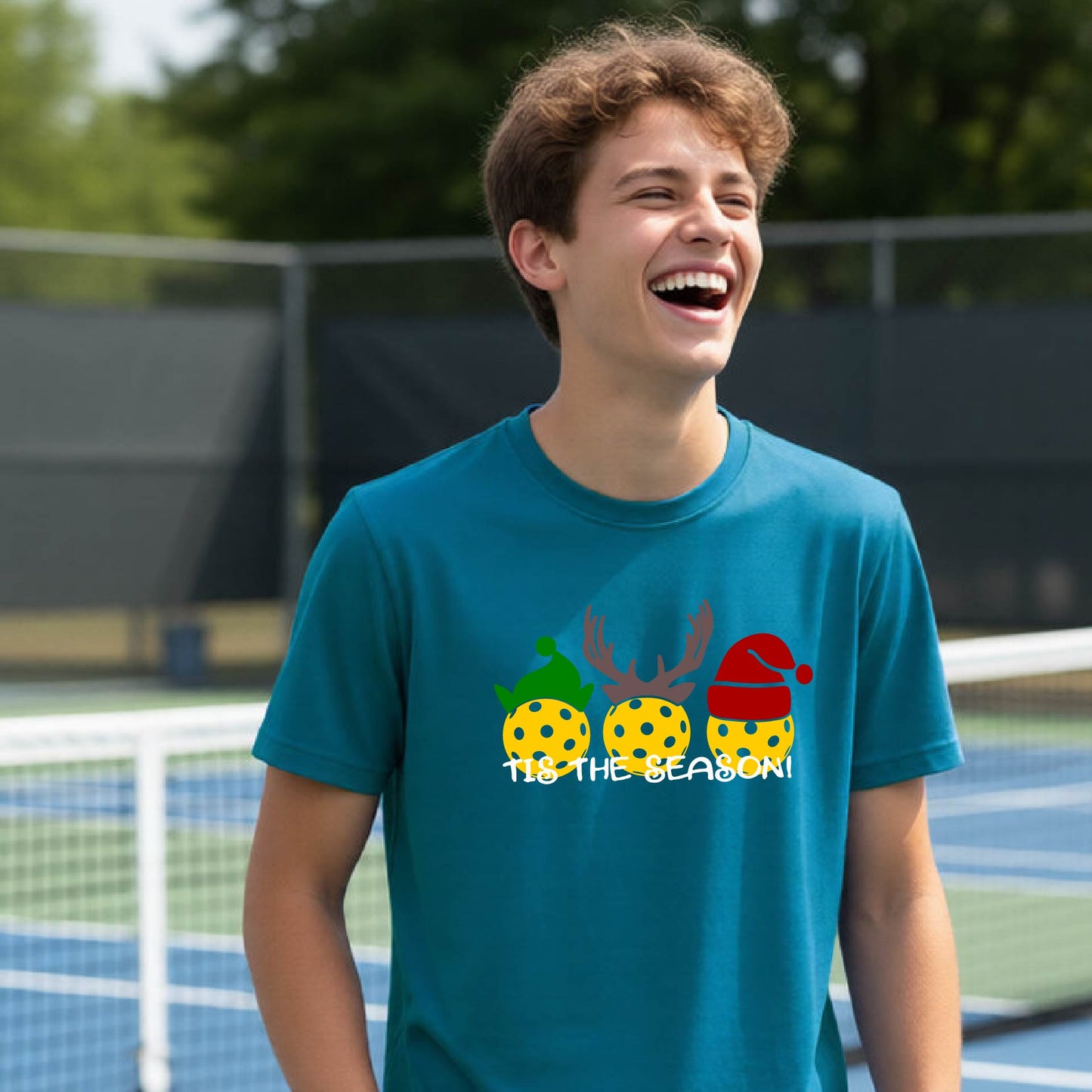 A young man laughing on an outdoor pickleball court, wearing a teal t-shirt with a festive graphic of three pickleball Christmas-themed golf balls and the text 'IT'S THE SEASON!'.