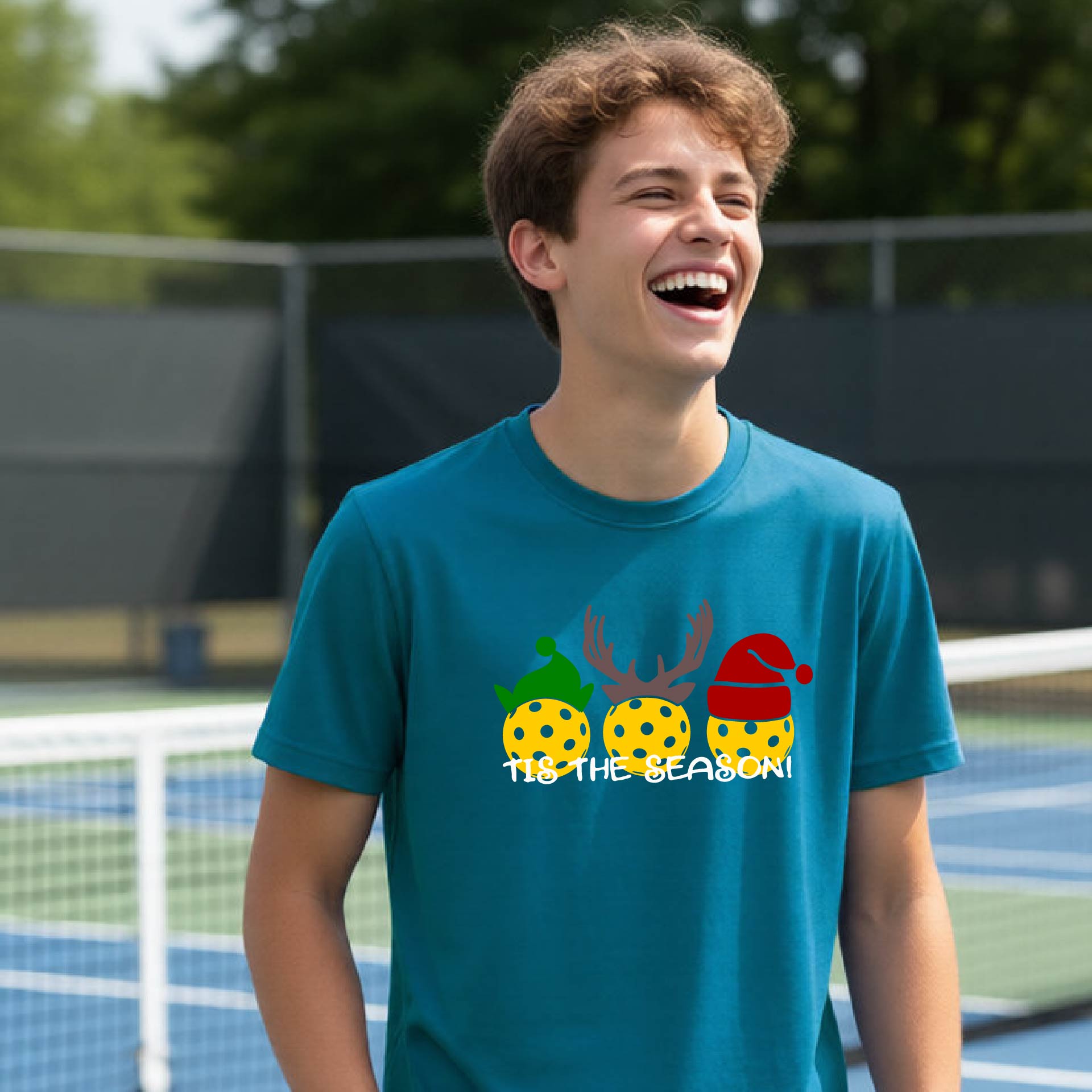A young man laughing on an outdoor pickleball court, wearing a teal t-shirt with a festive graphic of three pickleball Christmas-themed golf balls and the text 'IT'S THE SEASON!'.