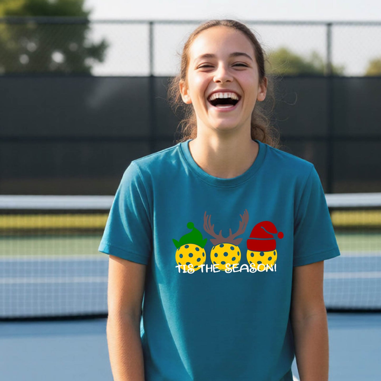 A young woman laughing on an outdoor pickleball court wearing a teal t-shirt with a festive graphic of three yellow polka-dot pickleballs decorated with green, brown, and red holiday accents, and the text 'TIS THE SEASON!'.