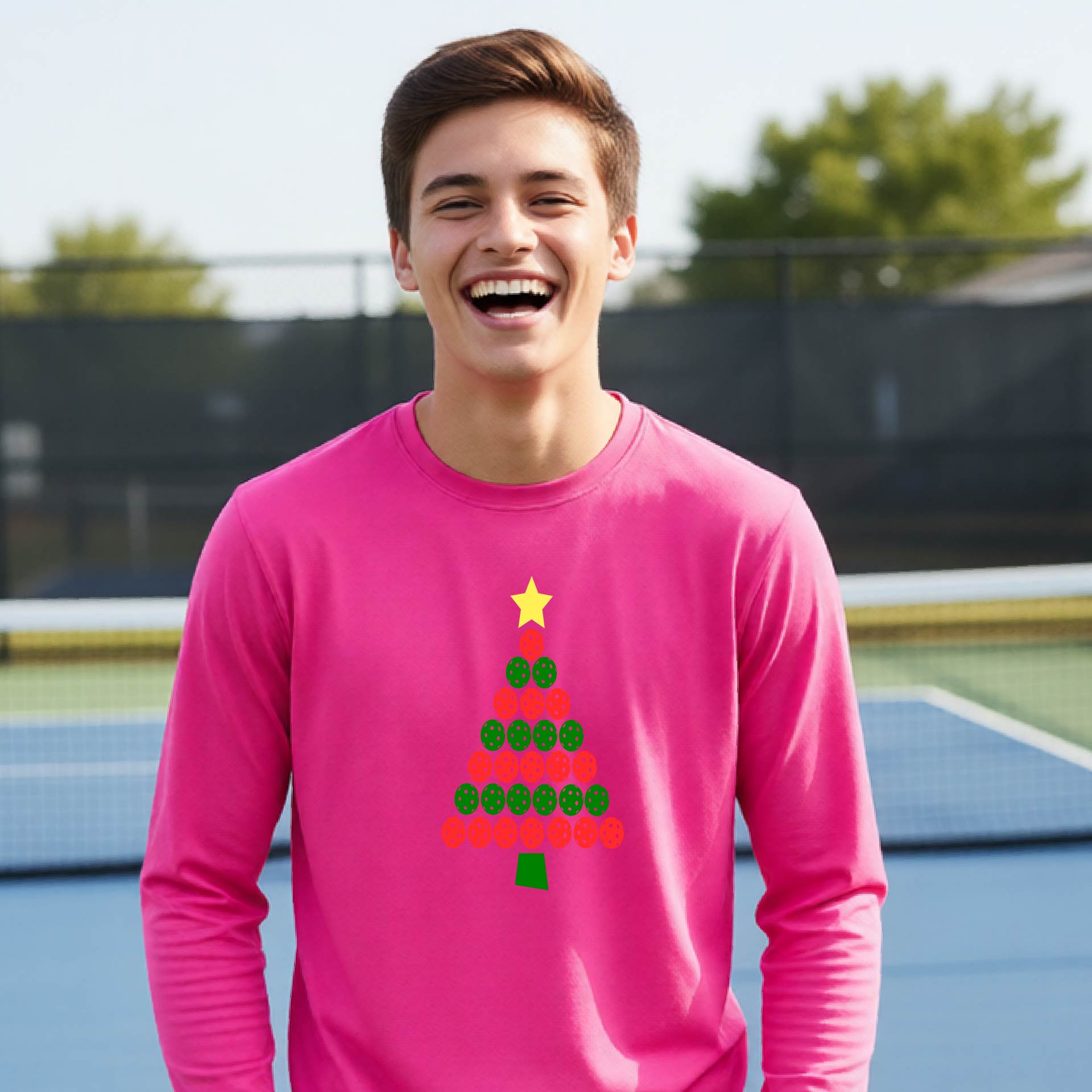 A young man smiling on an outdoor pickleball  court wearing a bright pink long-sleeve shirt with a Christmas tree graphic made of green and red pickleballs and a yellow star.