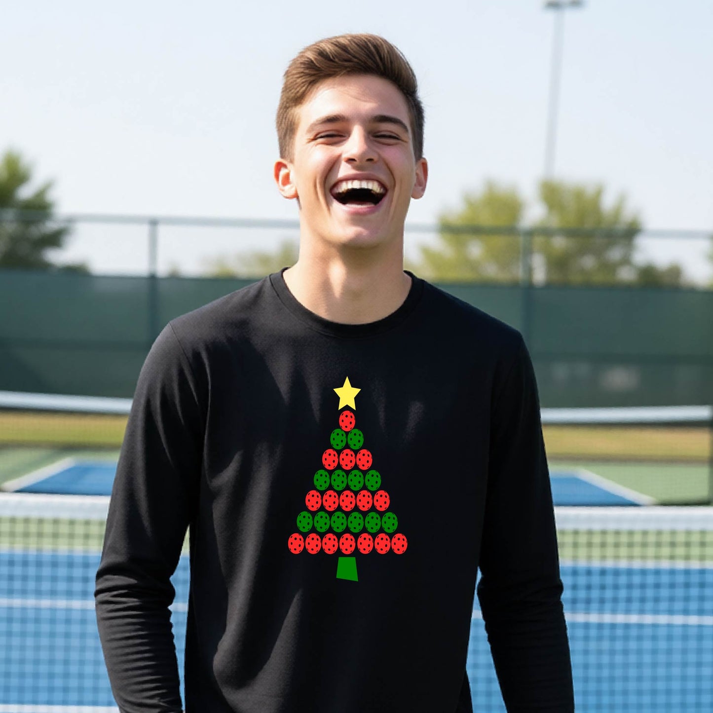 A young man laughing on an outdoor pickleball  court, wearing a black long-sleeve shirt with a Christmas tree graphic made of red and green pickleballs and a yellow star.