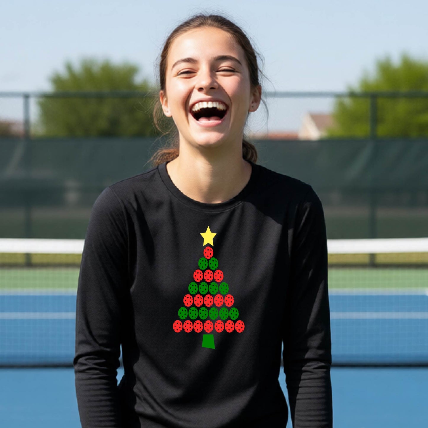 A woman laughing on an outdoor pickleball  court wearing a black long-sleeve shirt with a red and green Christmas tree graphic made of pickleballs on the front.