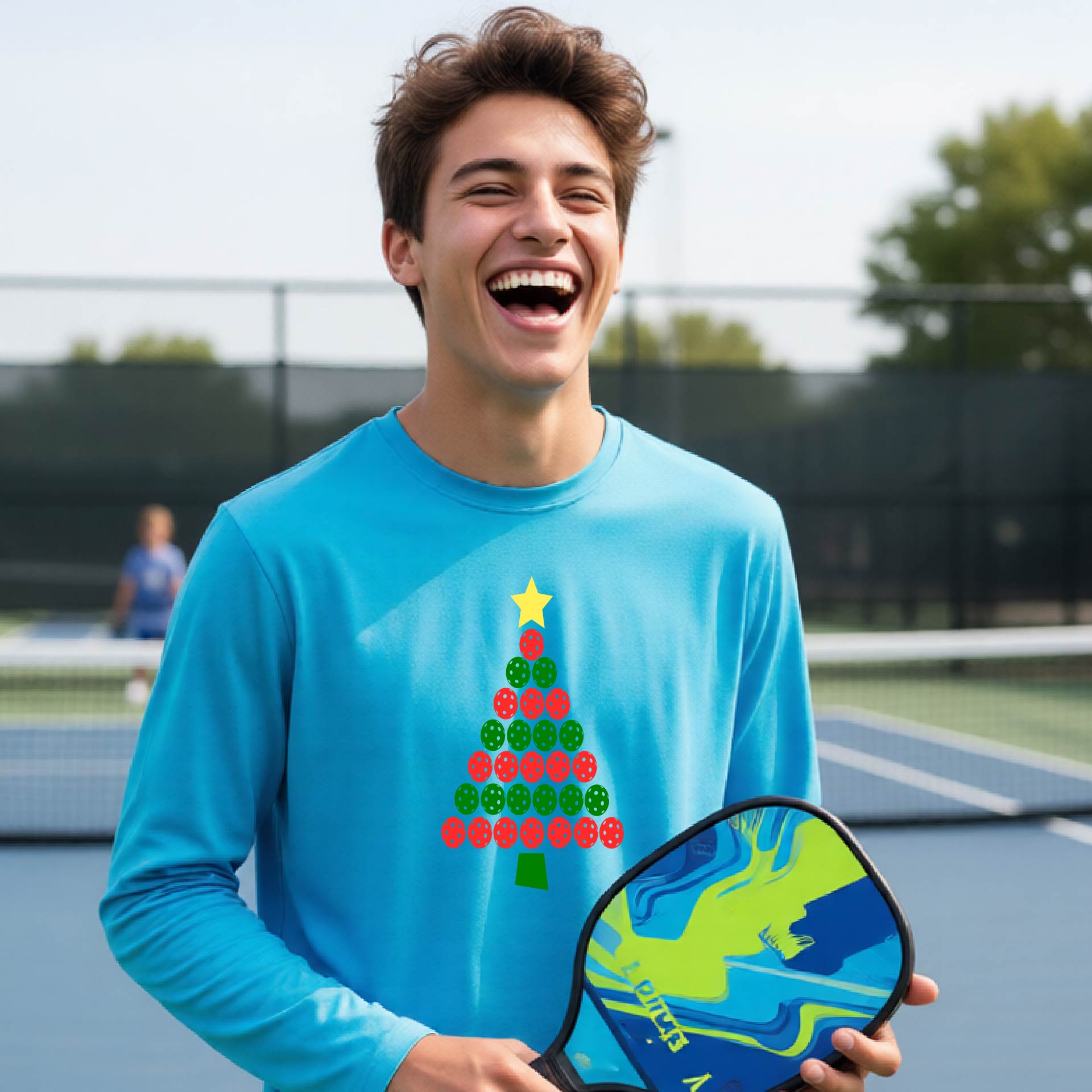 A man laughing on a pickleball court wearing a bright blue long-sleeve shirt with a Christmas tree made of pickleball balls on the front, holding a blue and green pickleball paddle.