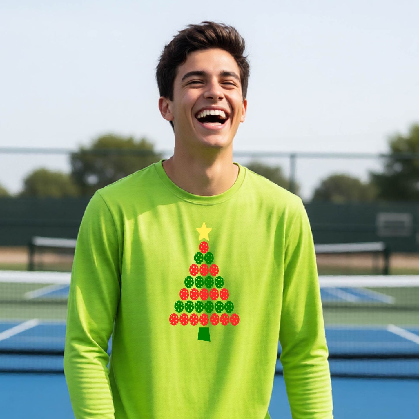 A man laughing on an outdoor pickleball  court wearing a bright lime green long-sleeve shirt with a Christmas tree made of pickleballs on the front.