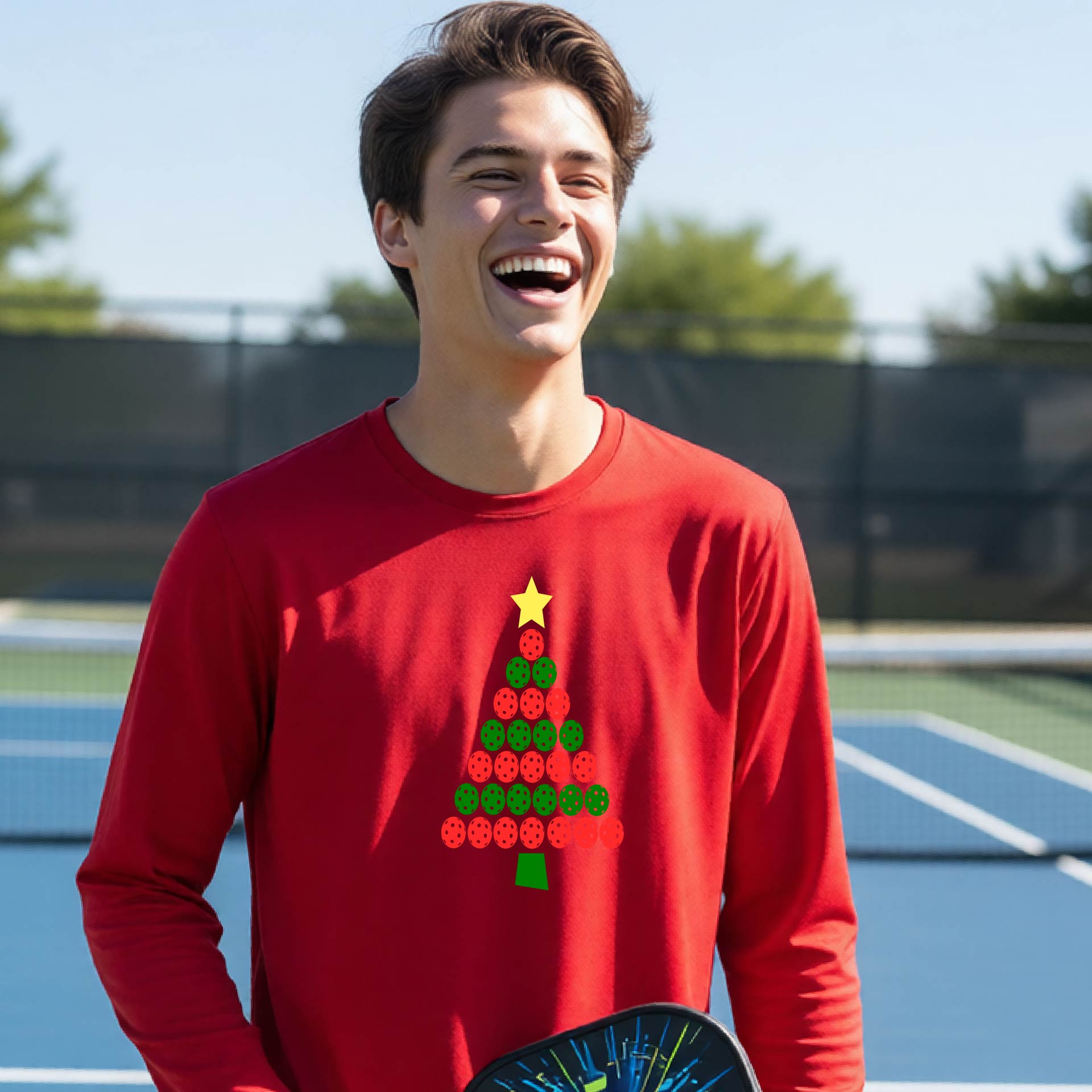A man laughing on an outdoor pickleball court wearing a red long-sleeve shirt with a Christmas tree made of red and green pickleballs and a yellow star on the chest.
