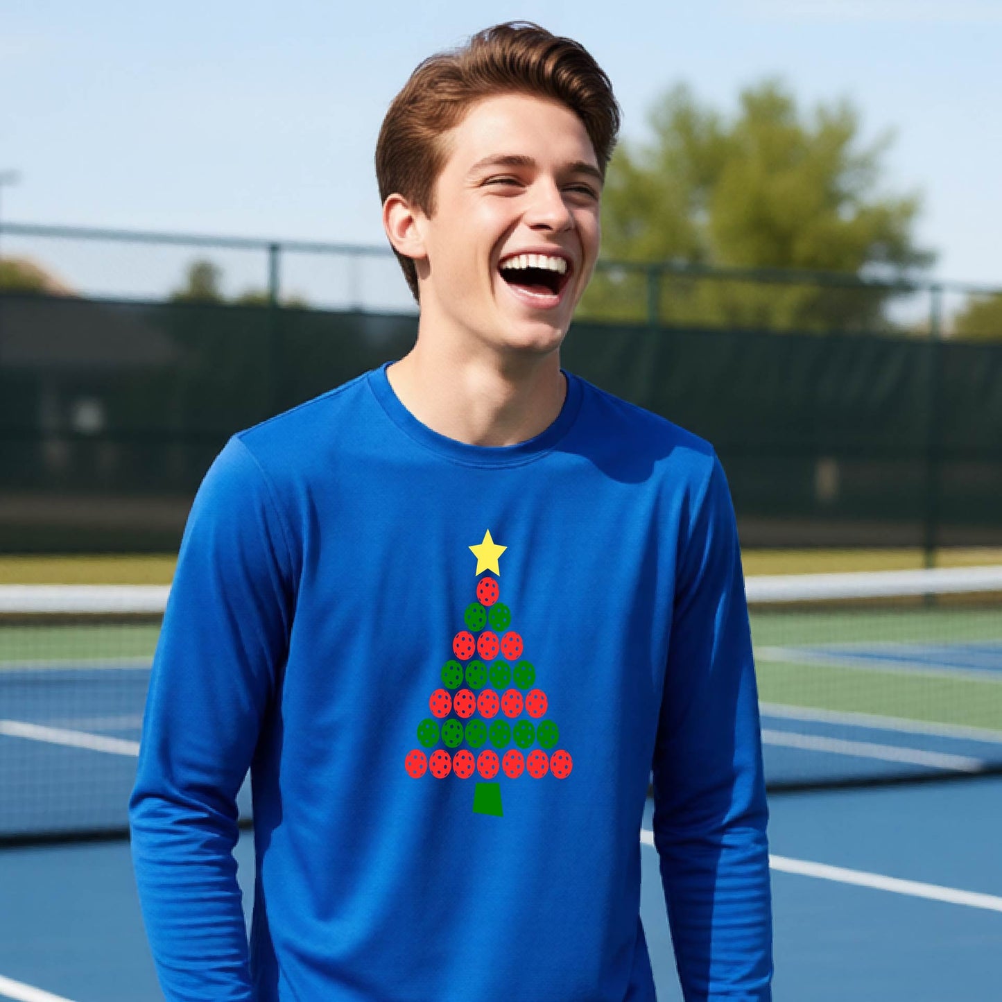 A man laughing on an outdoor pickleball  court wearing a bright blue long-sleeve shirt with a Christmas tree made of red and green pickleballs and a yellow star on the chest.